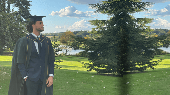 A man wearing an academic gown and cap stands outdoors, looking thoughtfully into the distance with a scenic background of trees and a lake.