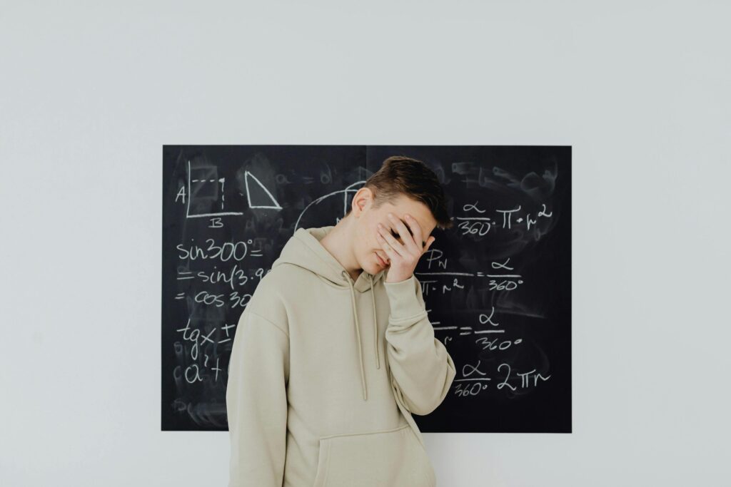 Teenager standing in front of a blackboard with math equations, covering his face in frustration.